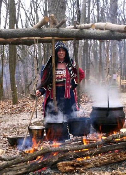An Ojibwa woman in traditional drss demonstarting how to boil maple syrup over an open fire using iron pots.